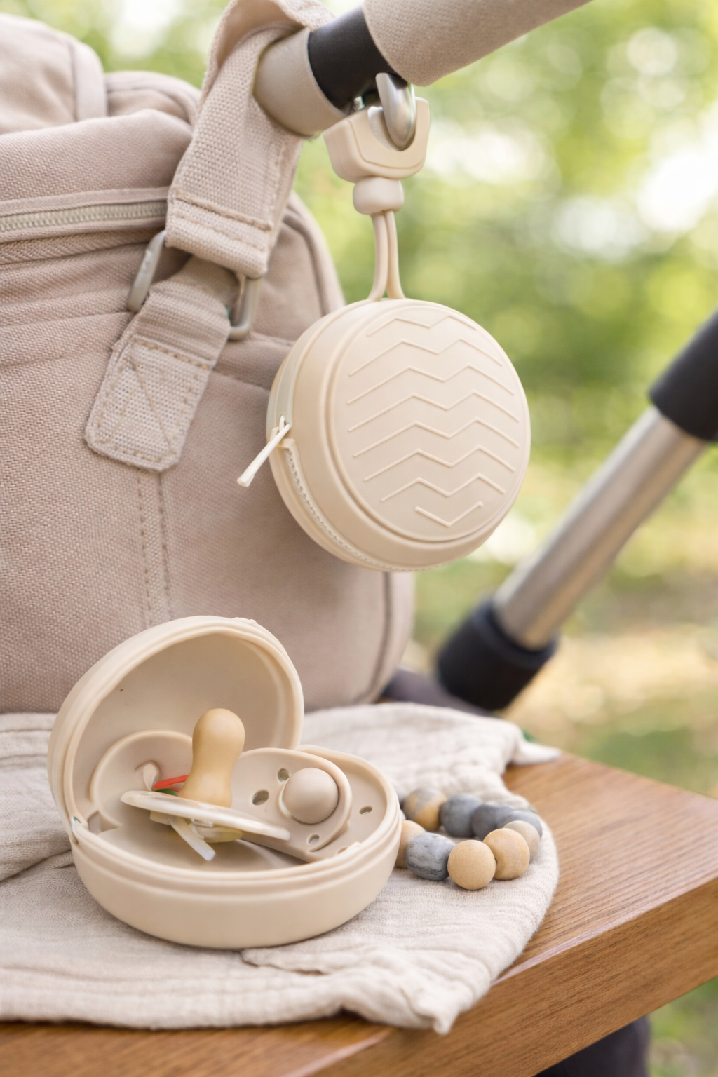 Cream silicone dummy holder clipped to a beige stroller bag, with a matching round pacifier case open on a wooden bench. The case contains a silicone pacifier and is styled with neutral-toned beads in a soft outdoor setting.