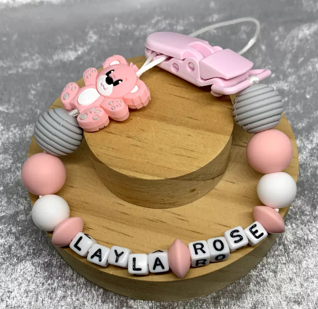Pink teddybear dummy holder on a grey textured surface. Personalised with the name 'Layla-Rose'. Change of name is available in the personalisation box. MAM Adapters are available too.