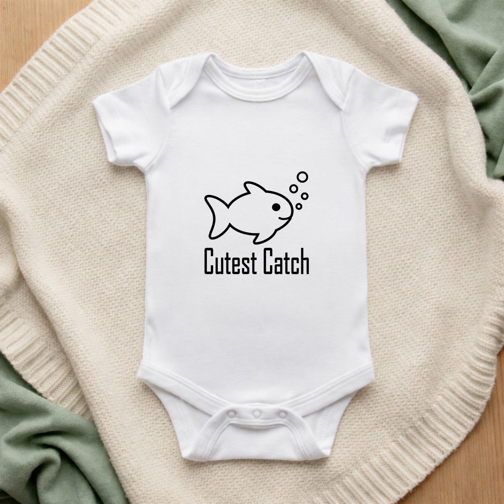 White baby onesie with fish graphic and 'Cutest Catch' text on a textured surface.