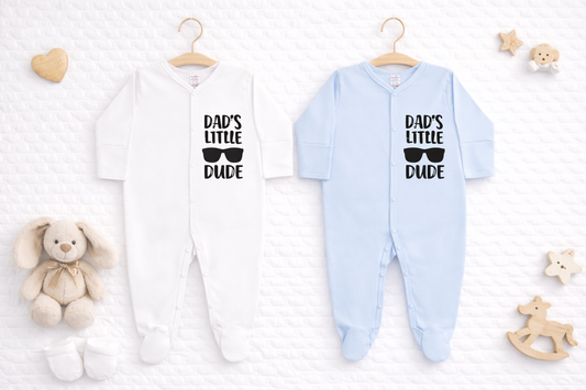 Mockup image showing two baby sleepsuits hanging on wooden hangers against a soft white quilted background. One sleepsuit is white and the other is light blue, both featuring black “Dad’s Little Dude” text with sunglasses graphic on the front. Styled with neutral baby props including a plush teddy, wooden heart, stars, rocking horse, and baby socks for a clean, modern product presentation.