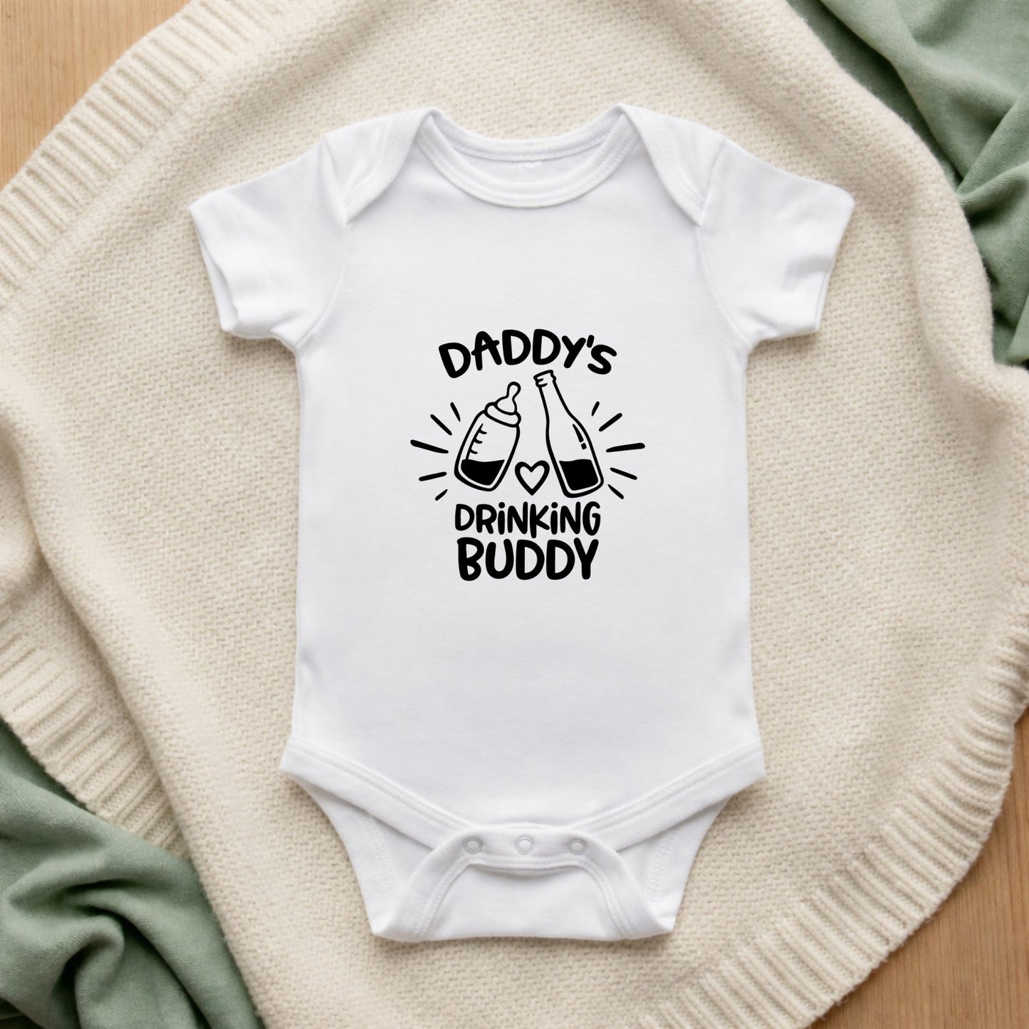 White baby onesie with 'Daddy's Drinking Buddy' text on a textured surface