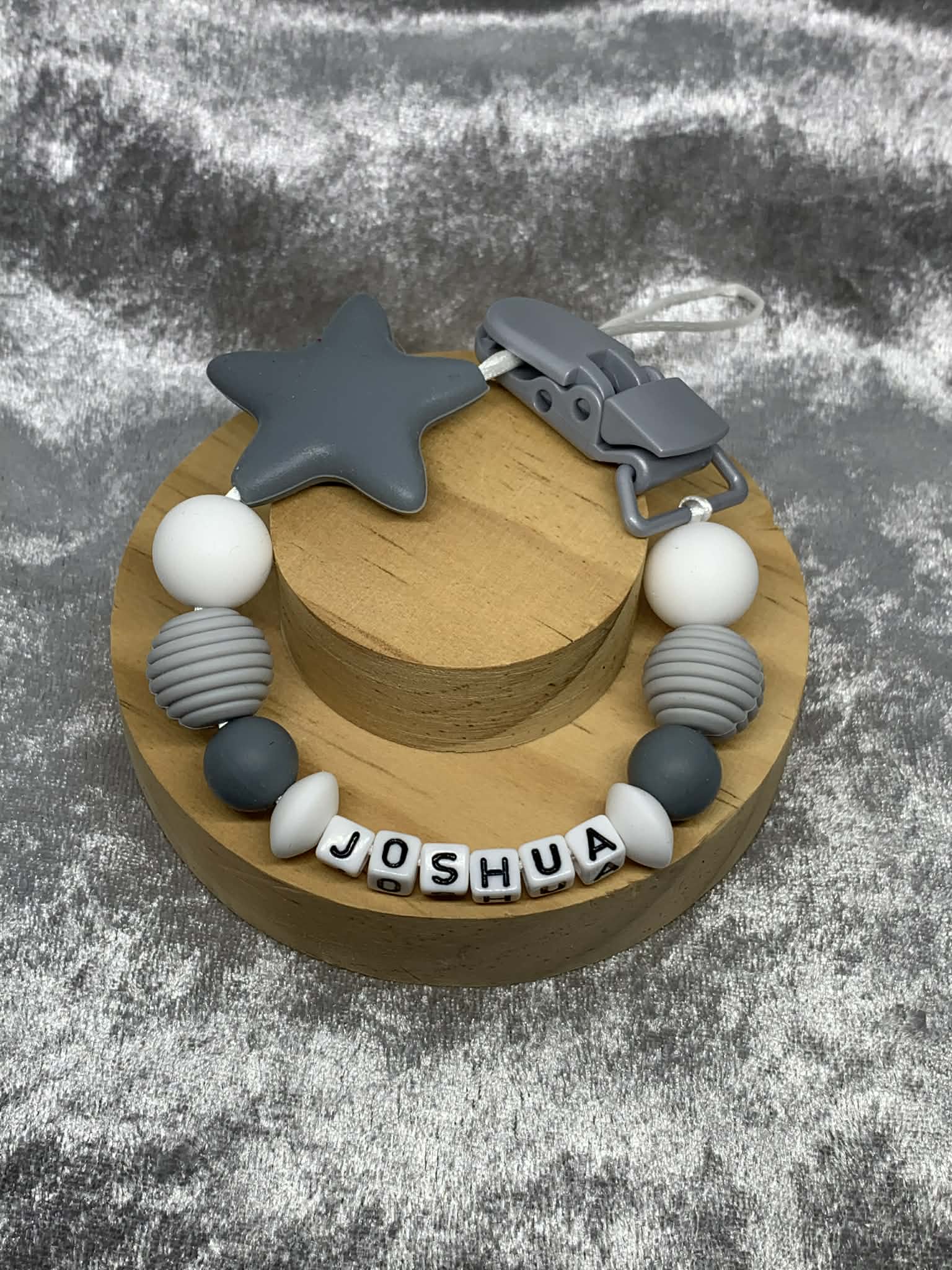 Dark Grey & White Silicone Dummy Clip with Dark Grey star personalised with the name 'Joshua' on a grey textured surface. Can be personalised