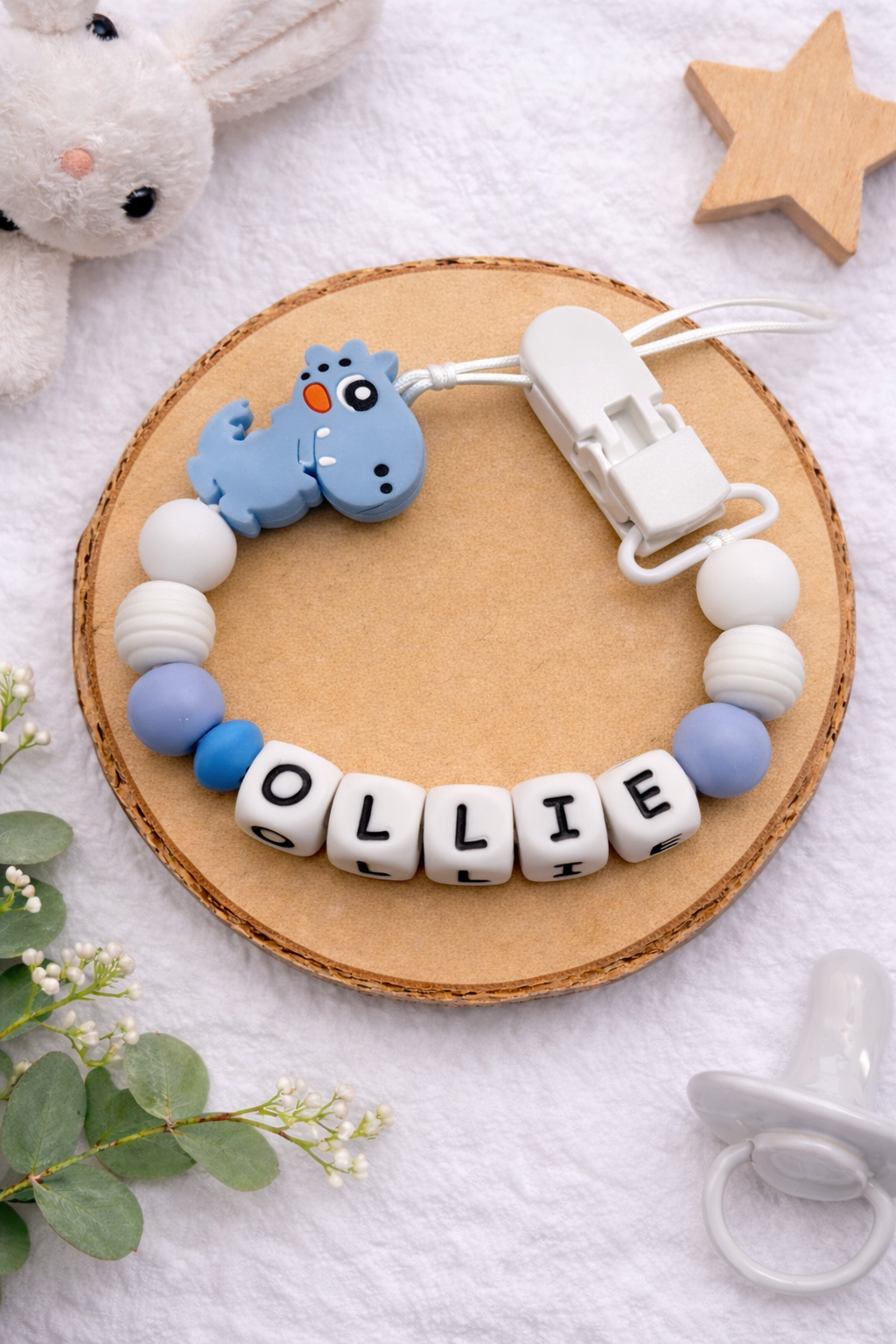 Flat lay of a blue and white dinosaur dummy clip with personalised name.