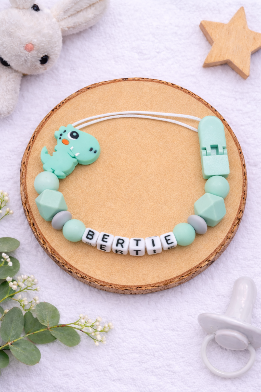 Flat lay of a green and grey dinosaur dummy clip with personalised name.