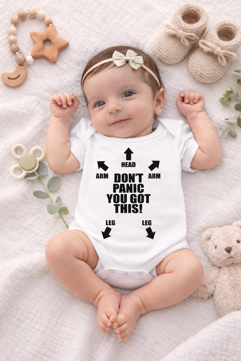 Baby wearing a white onesie with text, surrounded by toys and baby items on a soft surface.