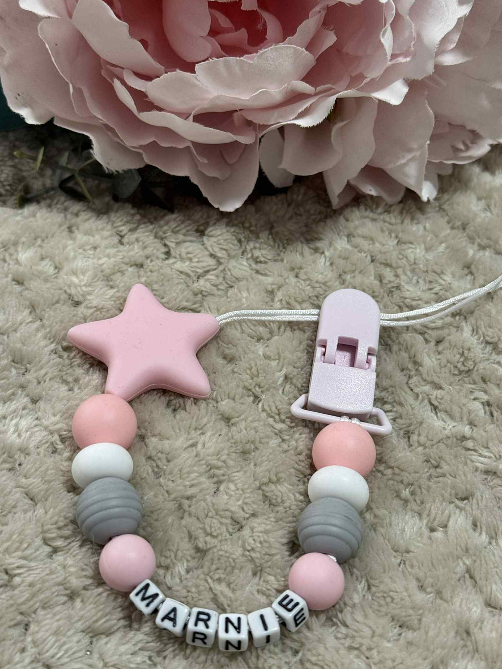 A beaded pacifier clip with pink, White and grey beads, a pink star, and letter beads spelling "Marnie" lying on a textured Beige surface. Personalised option available