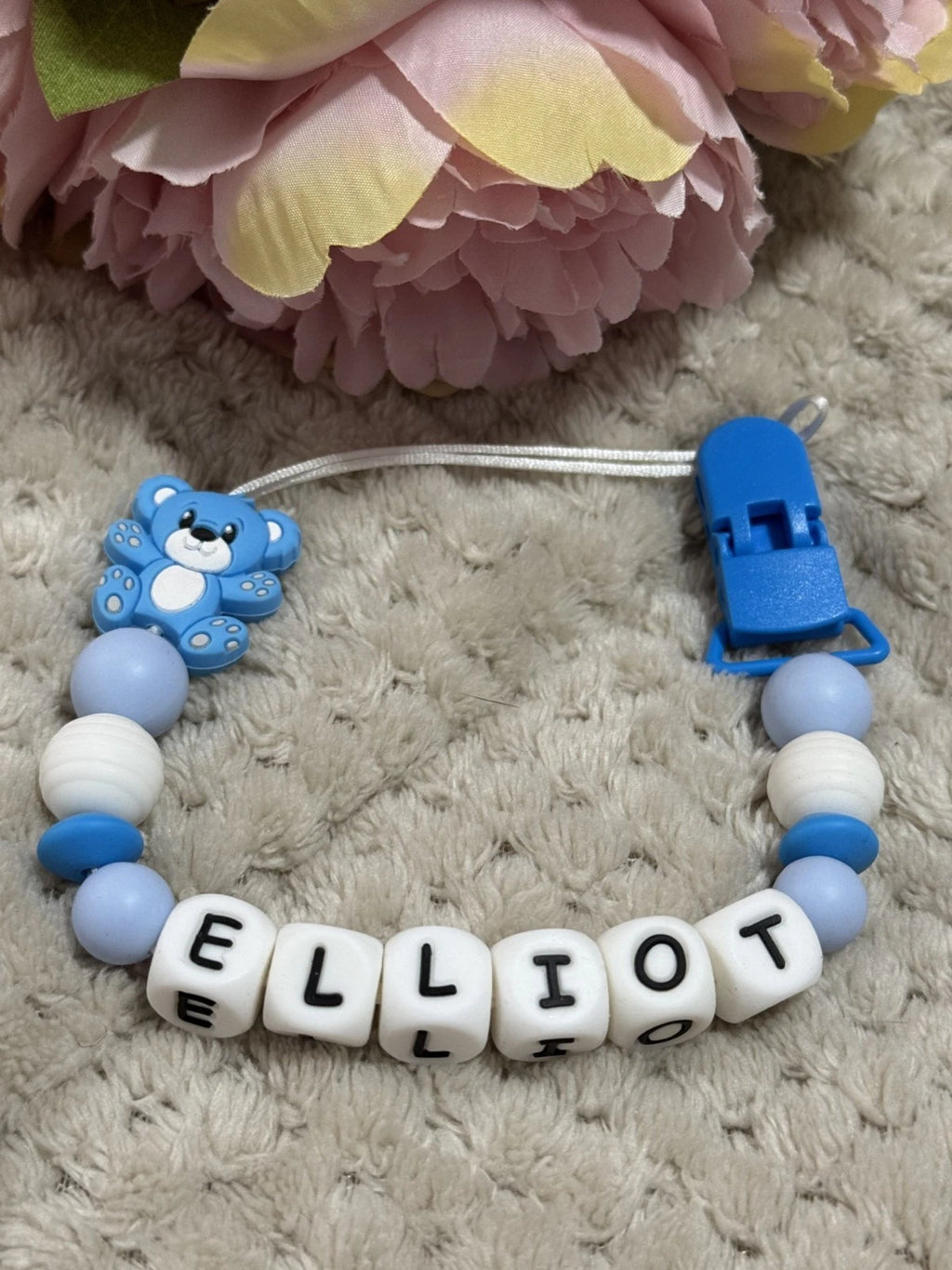 Dummy Clip with letter beads spelling 'Elliot' on a textured surface with flowers in the background. Personalisation and MAM Adapters available.