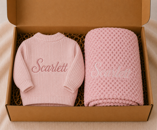 Pink baby sweater and blanket with 'Scarlett' embroidery in a cardboard box.