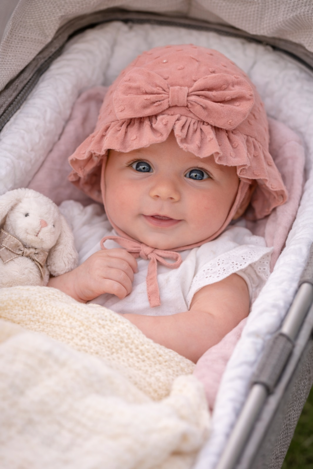 Baby in a pink bonnet sitting in a crib with a teddy bear.