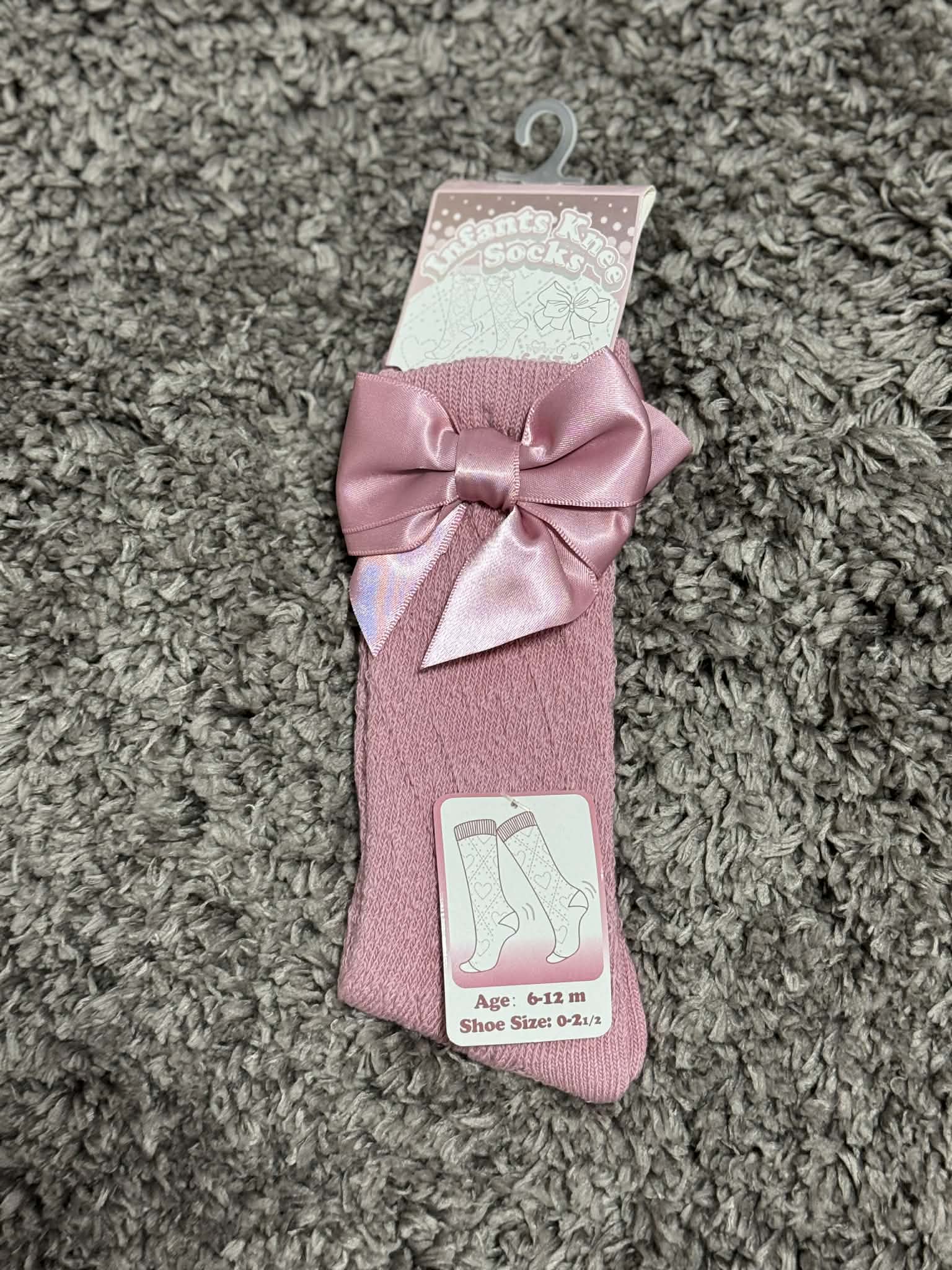 Pink baby socks with a bow on a hanger against a carpeted floor.