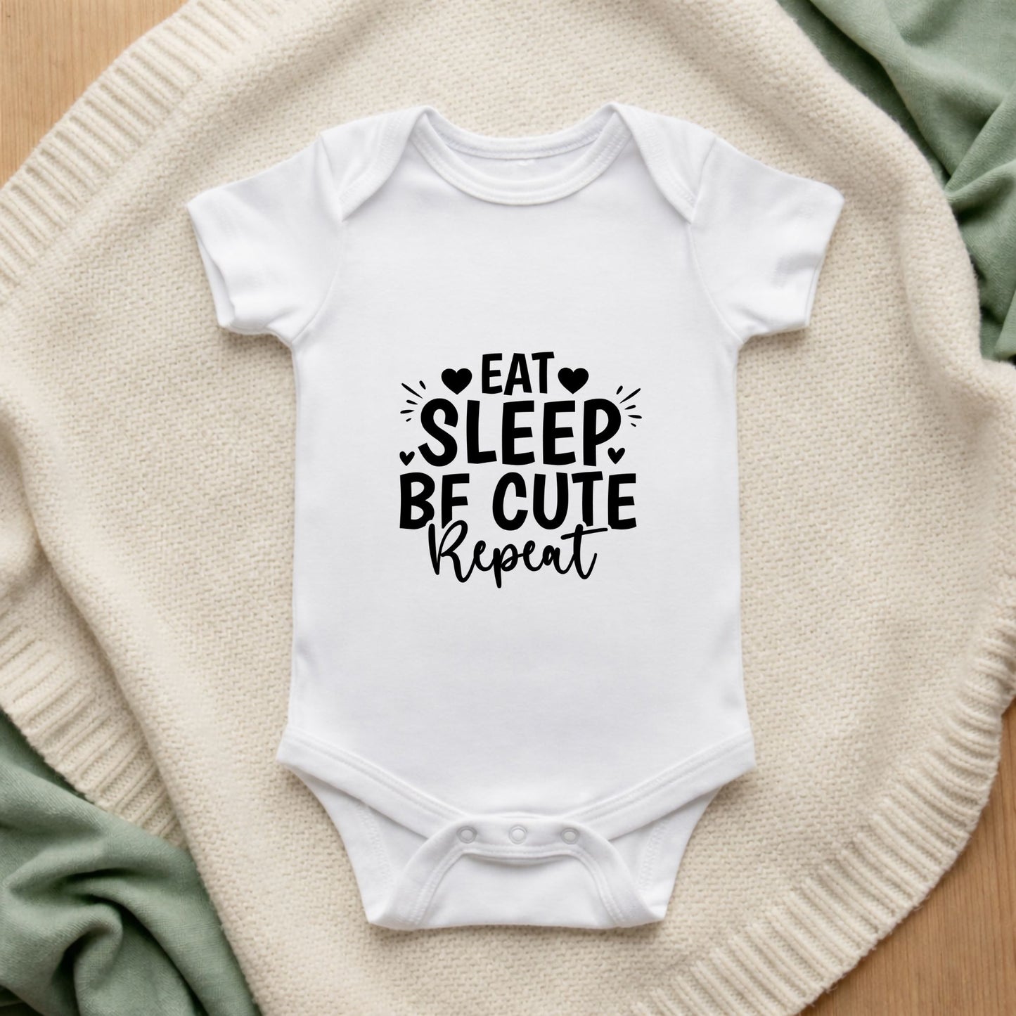 White baby onesie with 'Eat Sleep Be Cute Repeat' text on a textured surface.