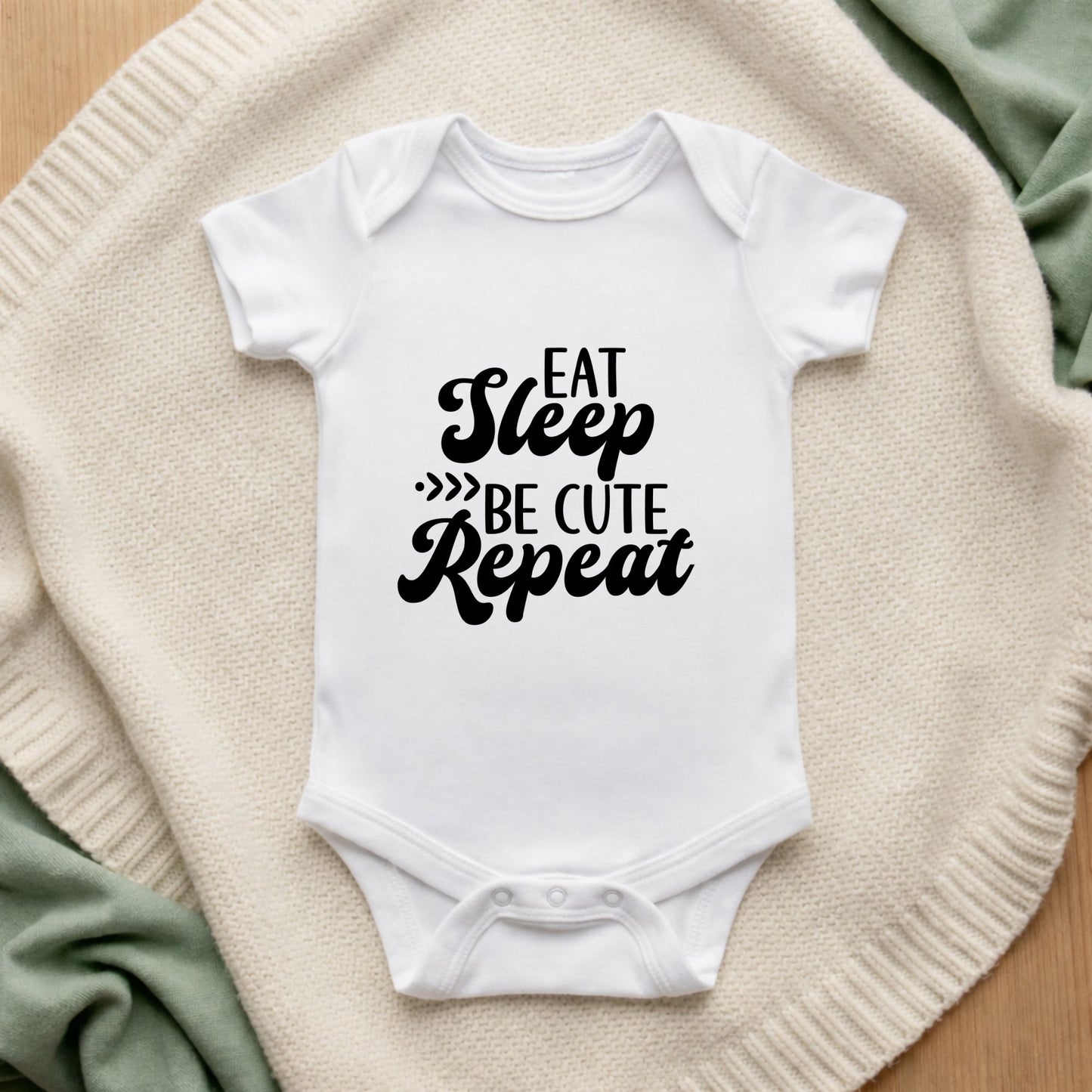 White baby onesie with 'Eat Sleep Be Cute Repeat' text on a textured surface.