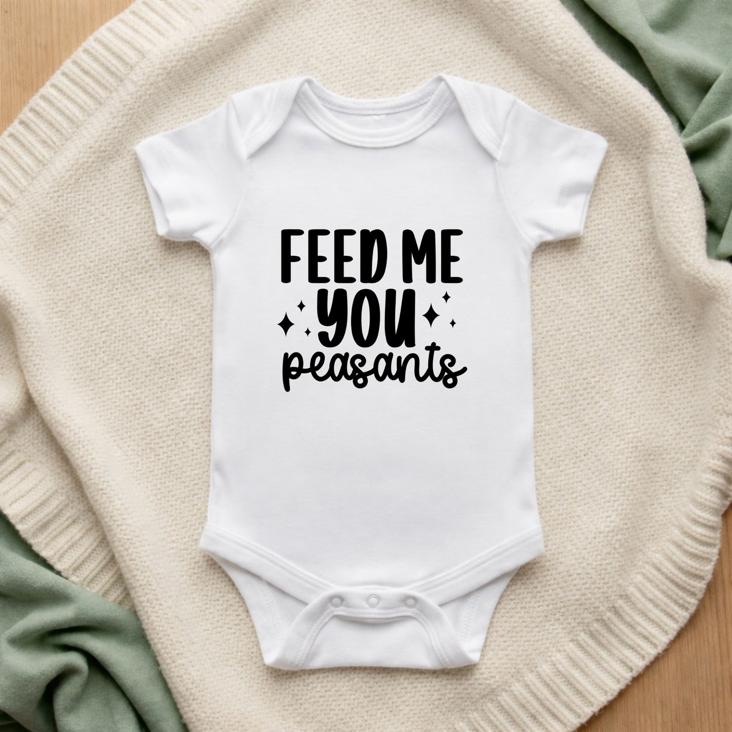 White baby onesie with 'FEED ME YOU PEASANTS' text on a textured surface.