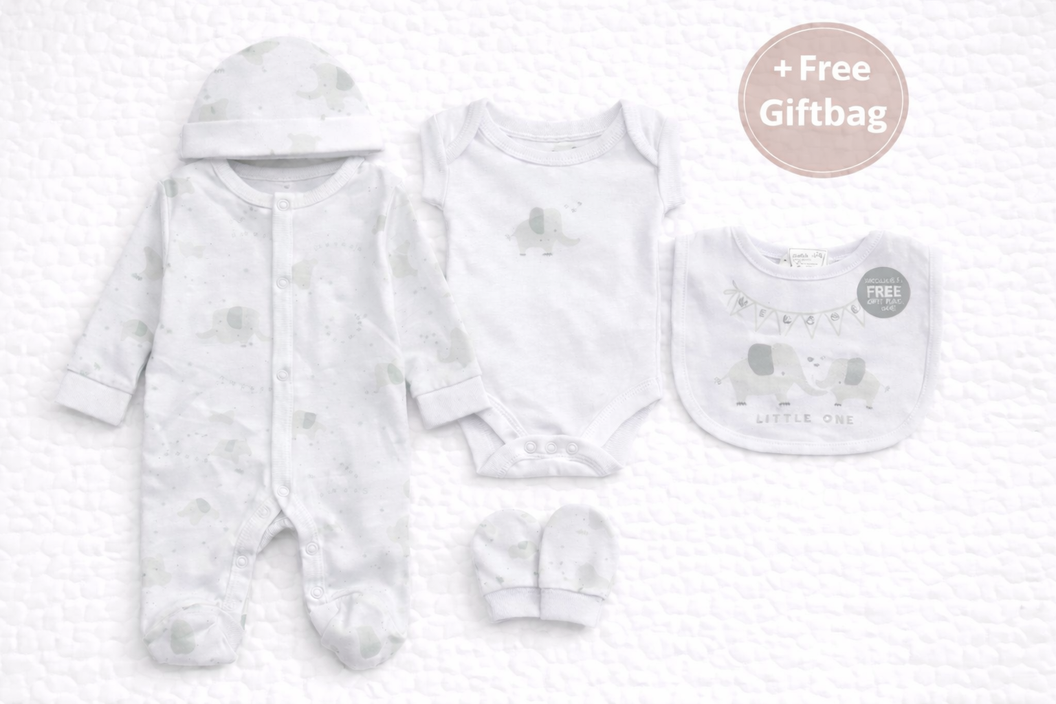 Flat lay mockup of a five-piece baby clothing gift set displayed on a white quilted background. The set includes a white elephant-print footed sleepsuit, short-sleeve bodysuit, matching hat, mittens, and coordinating bib featuring soft grey and sage elephant designs. A circular “+ Free Giftbag” label is shown, with no organza bag visible.