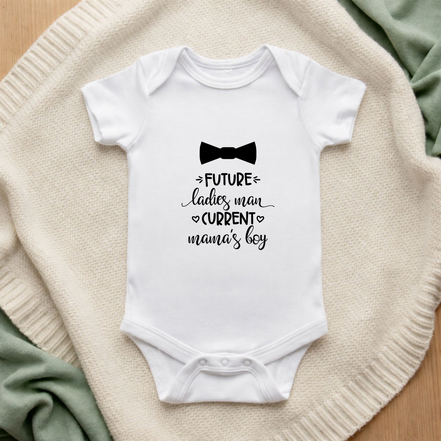 White baby onesie with text and a bow tie graphic on a textured surface. Future Ladies Man Current Mama's Boy Baby Vest