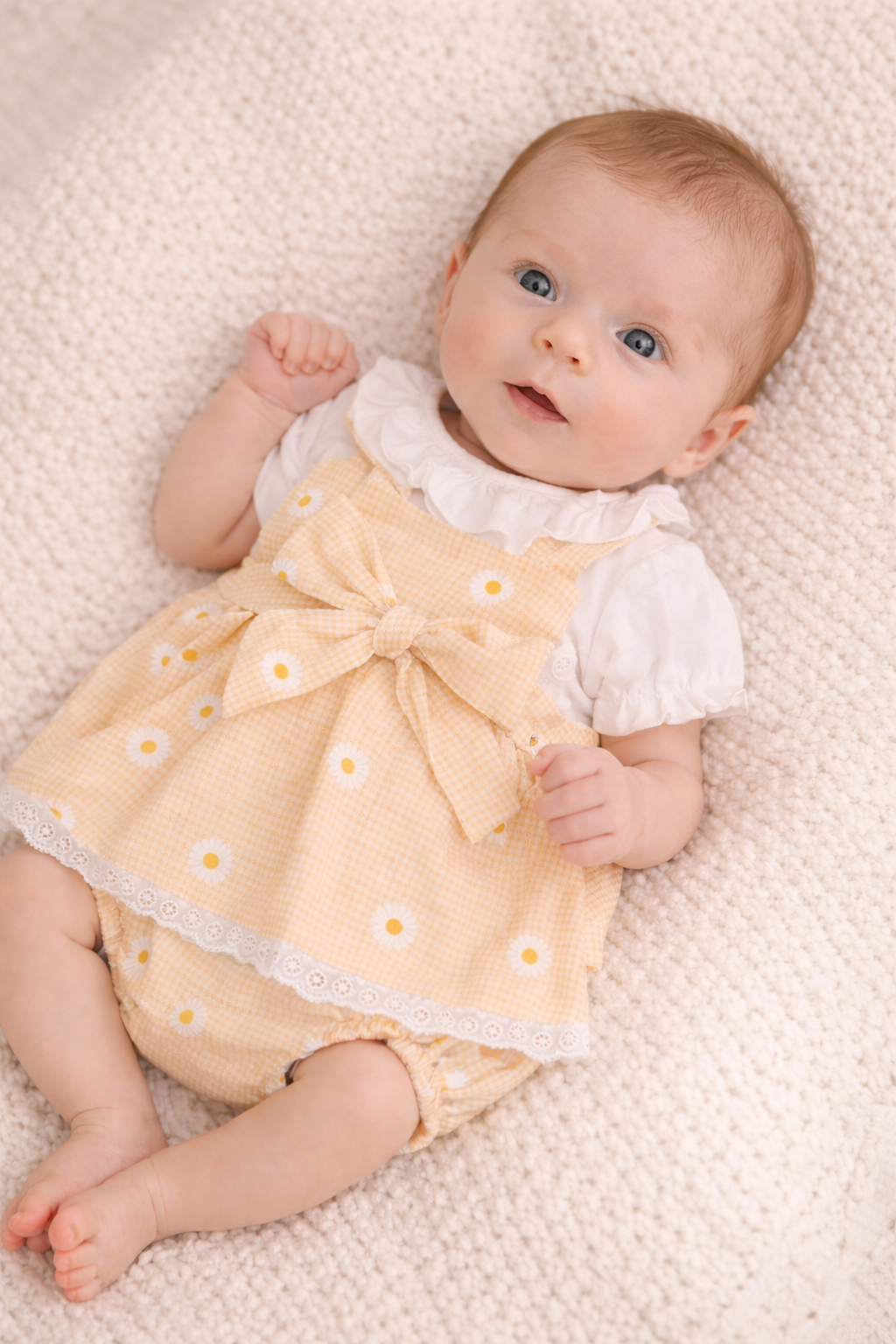 Baby wearing a yellow dress with white flowers on a beige blanket