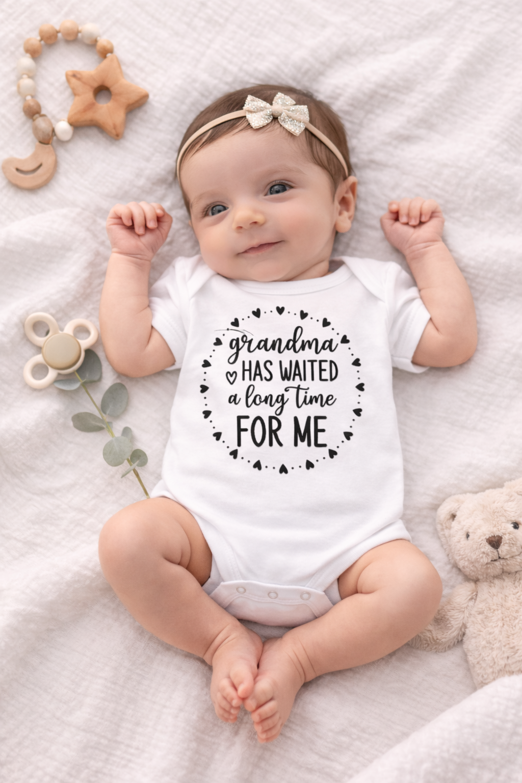 Baby wearing a onesie with 'Grandma has waited a long time for me' text, surrounded by toys on a soft surface.