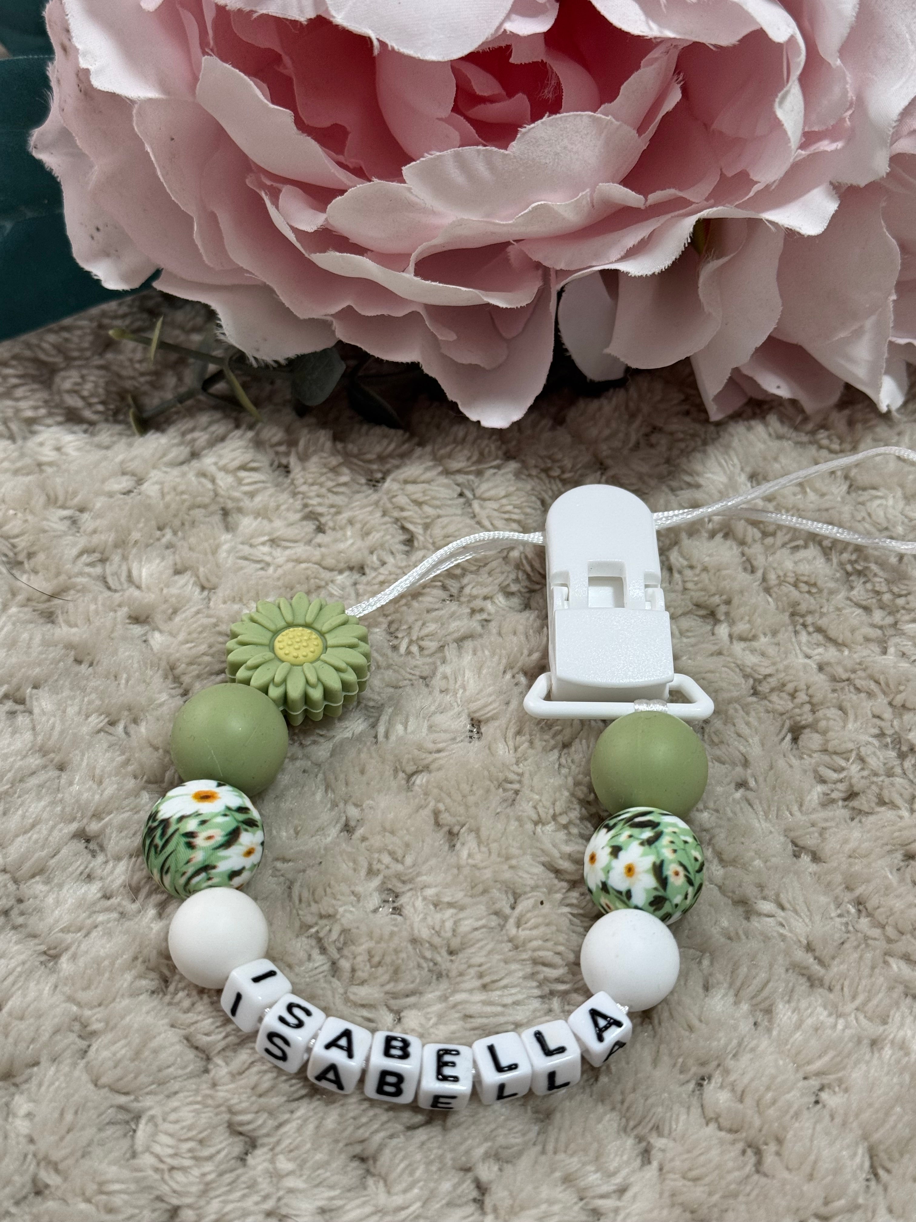 Baby dummy clip with colorful beads and a name tag next to a Green flower on a textured surface
