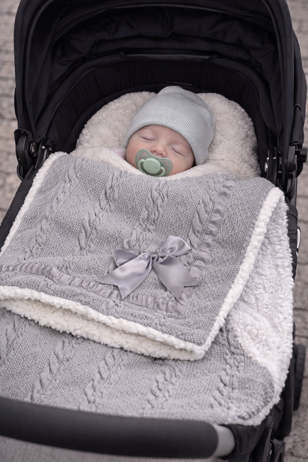 Sleeping newborn baby in a pram wrapped in a soft grey cable knit baby blanket with white sherpa fleece backing and decorative satin bow.