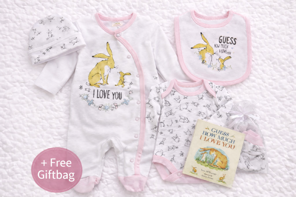 Guess How Much I Love You themed baby gift set featuring a white babygrow with all-white sleeves, a long-sleeved bodysuit with delicate bunny print, matching bib with pink trim, soft hat, scratch mittens, and a mini storybook. Beautifully coordinated in soft white and blush tones and presented with a free gift bag, perfect for newborn gifting.