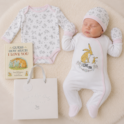 Newborn baby in a white onesie with bunny design, surrounded by baby clothes and a book on a beige surface.