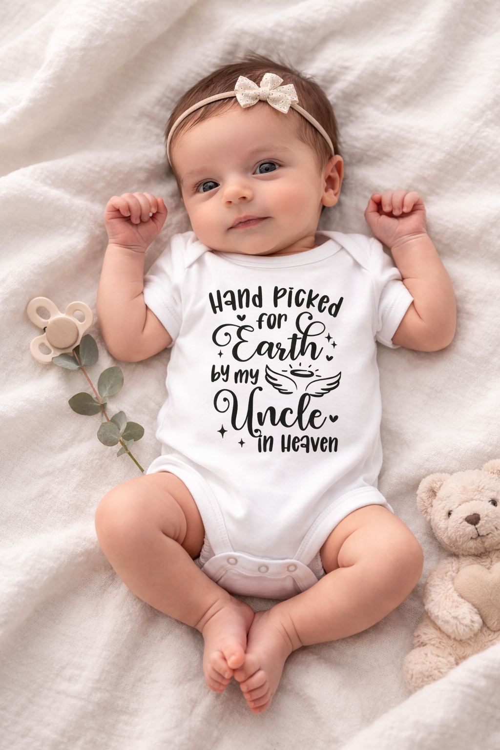 Baby wearing a onesie with text, lying on a soft surface with a teddy bear and plant.