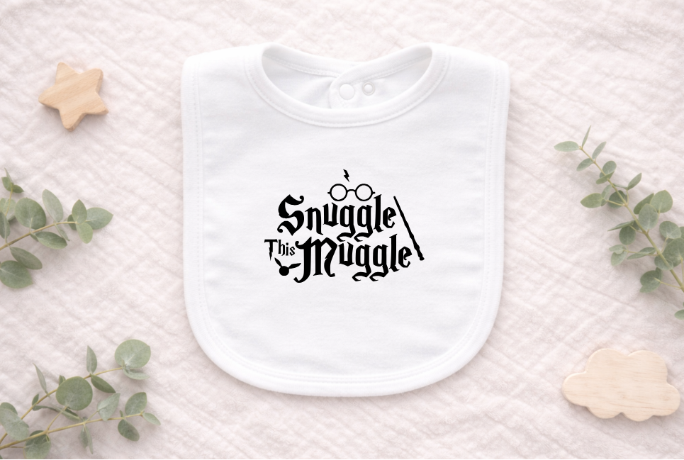 Flat lay of a white newborn baby bib with “Snuggle This Muggle” print, styled with soft neutral props for a cosy, whimsical baby gift presentation.
