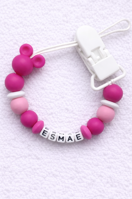 Pink and white baby Dummy Clip with 'ESMAE' name tag on a light purple background