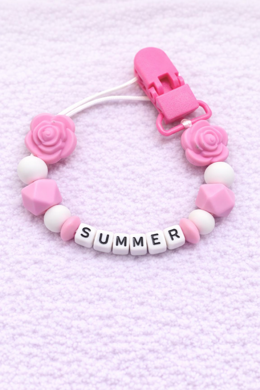 Pink and white pacifier clip with 'SUMMER' letter beads on a pink textured background