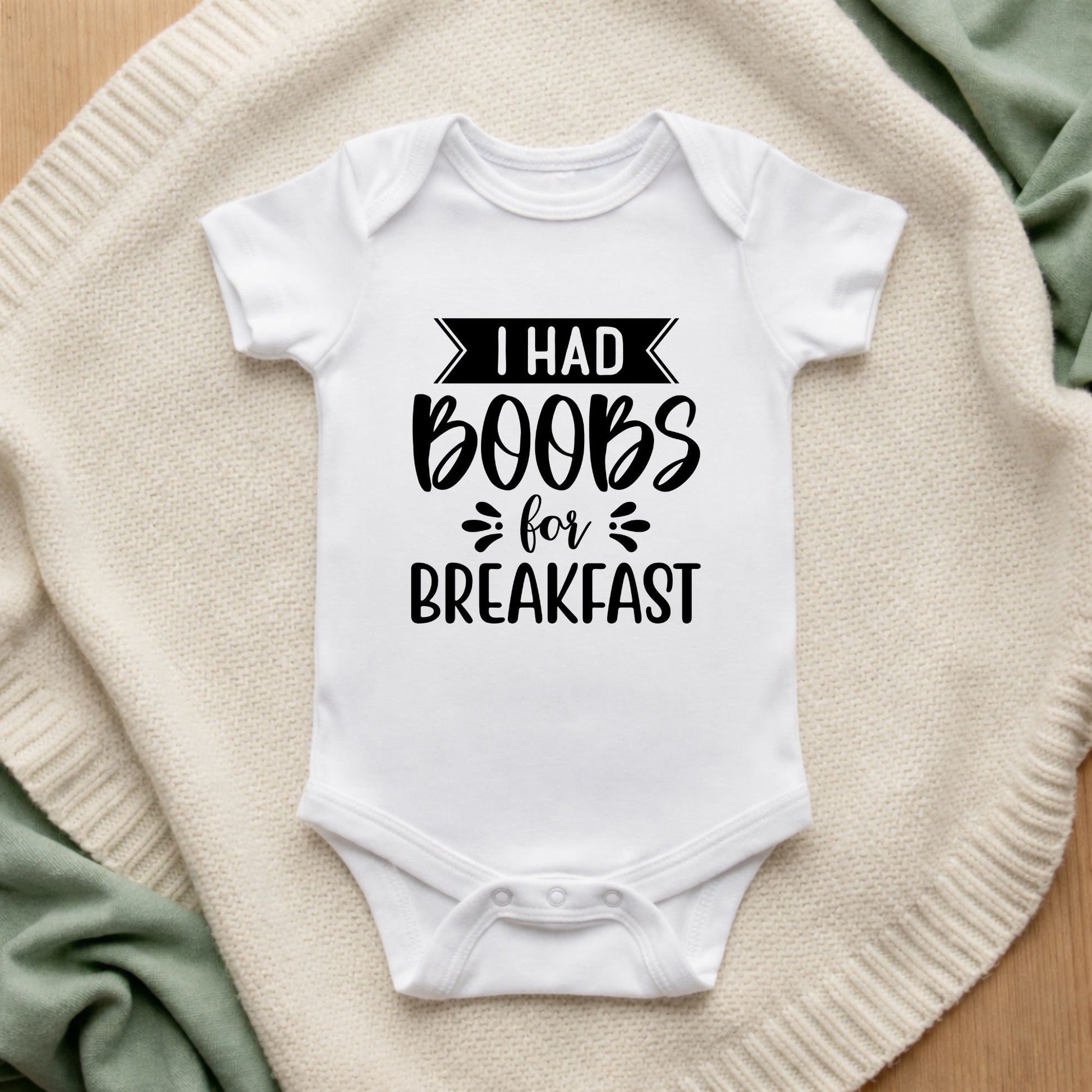 White baby onesie with text 'I had boobs for breakfast' on a textured surface.