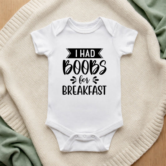 White baby onesie with text 'I had boobs for breakfast' on a textured surface.