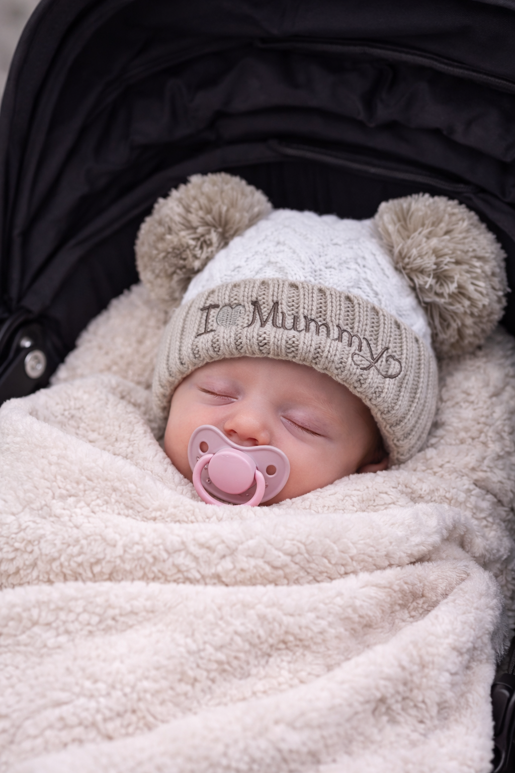 Sleeping newborn baby wearing a coffee brown and cream knitted hat with double pom poms and “I Love Mummy” embroidered detail, swaddled in a soft neutral blanket for a cosy newborn photoshoot mock-up.
