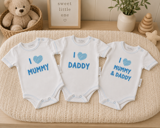 Pack of 3 white baby bodysuits with I Love Mummy, I Love Daddy and I Love Mummy & Daddy blue slogans, laid flat on a cream knit surface