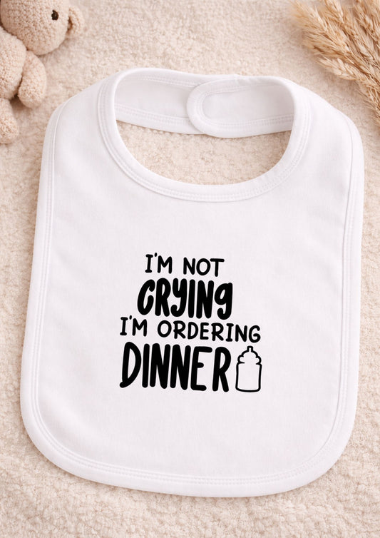 White baby bib with text 'I'm not crying, I'm ordering dinner' on a beige surface.
