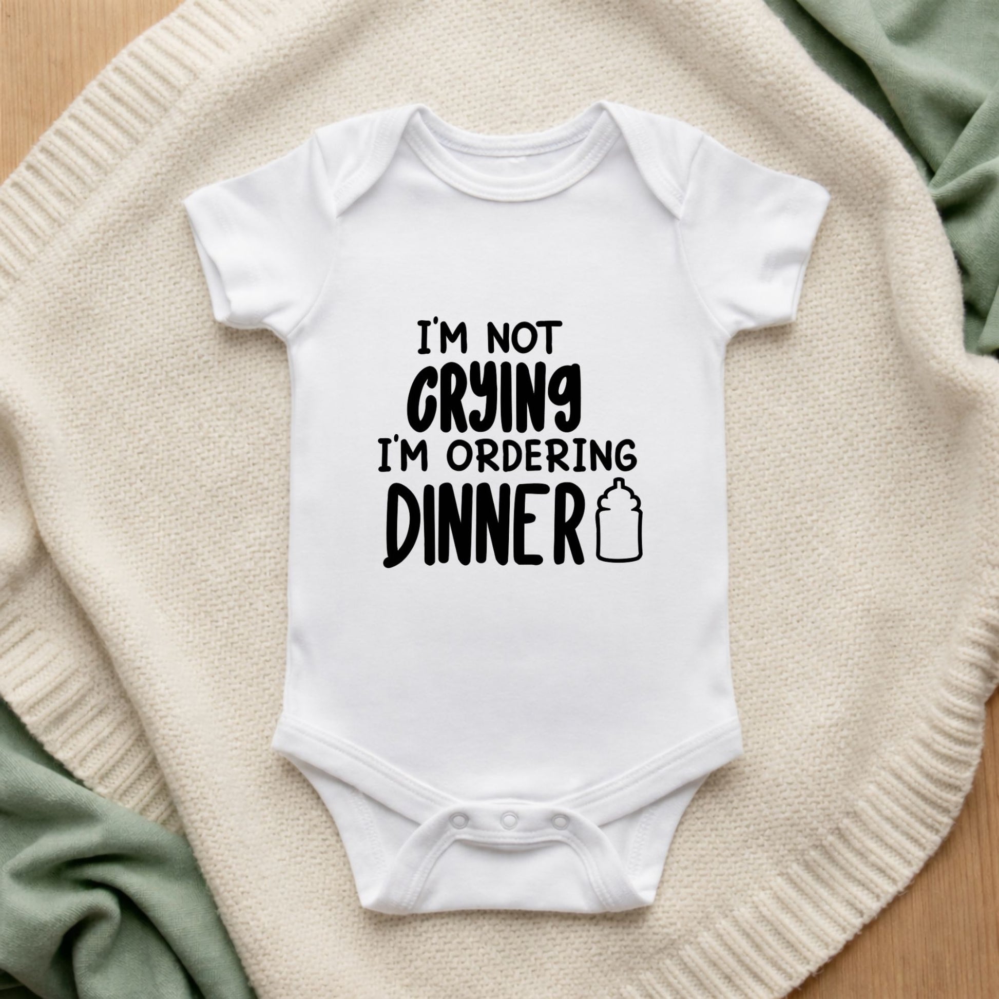 White baby onesie with text 'I'm not crying, I'm ordering dinner' on a textured surface.
