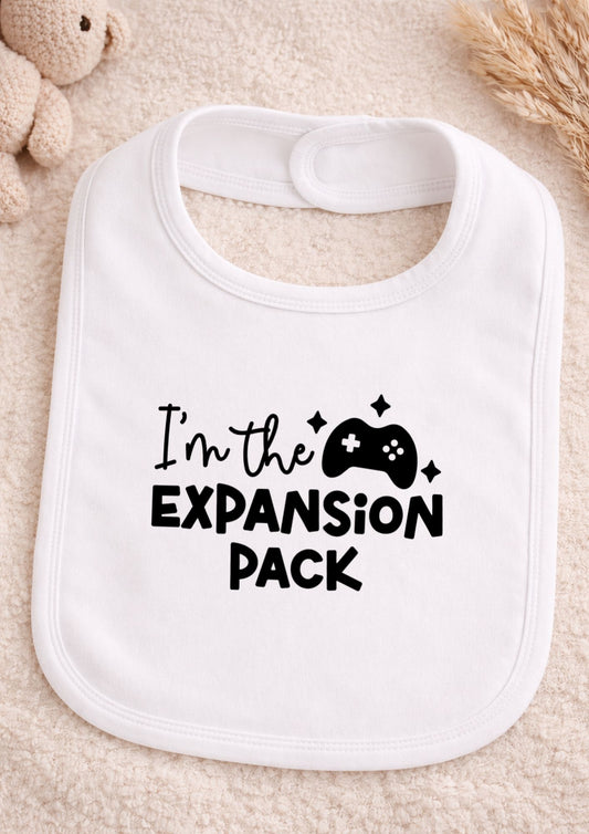 White baby bib with text 'I'm the Expansion Pack' on a beige surface