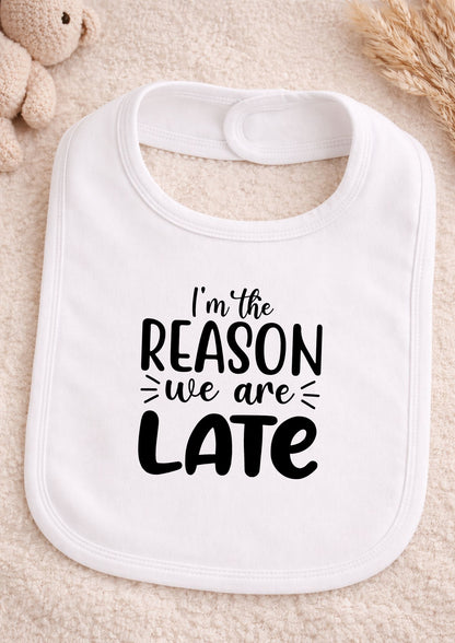 White baby bib with text 'I'm the reason we are late' on a beige surface.