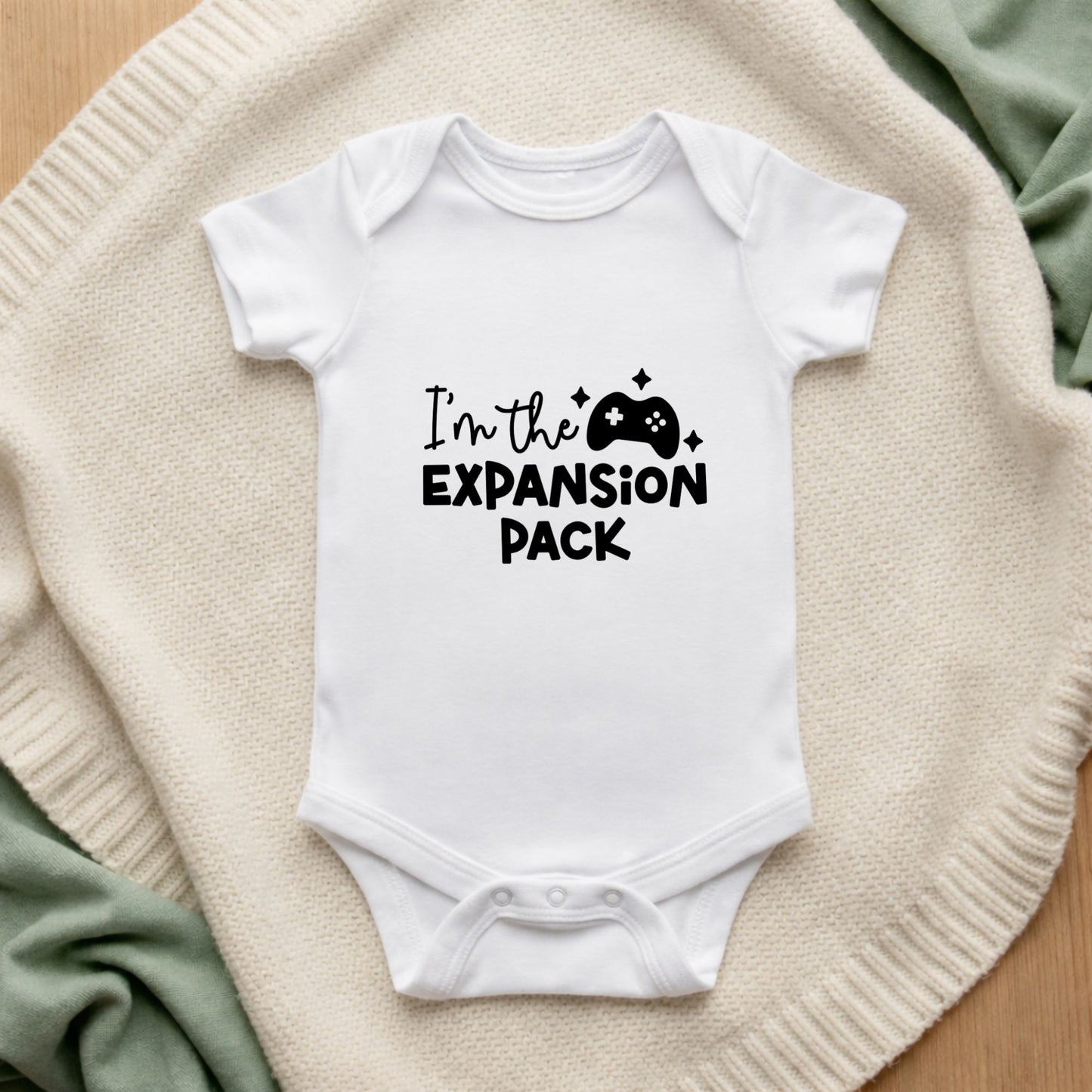 White baby onesie with 'I'm the Expansion Pack' text on a beige blanket