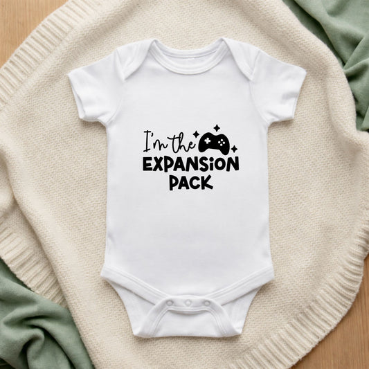 White baby onesie with 'I'm the Expansion Pack' text on a beige blanket