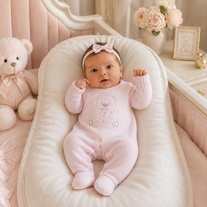Baby in a pink outfit with a teddy bear design, sitting on a white cushion in a nursery.