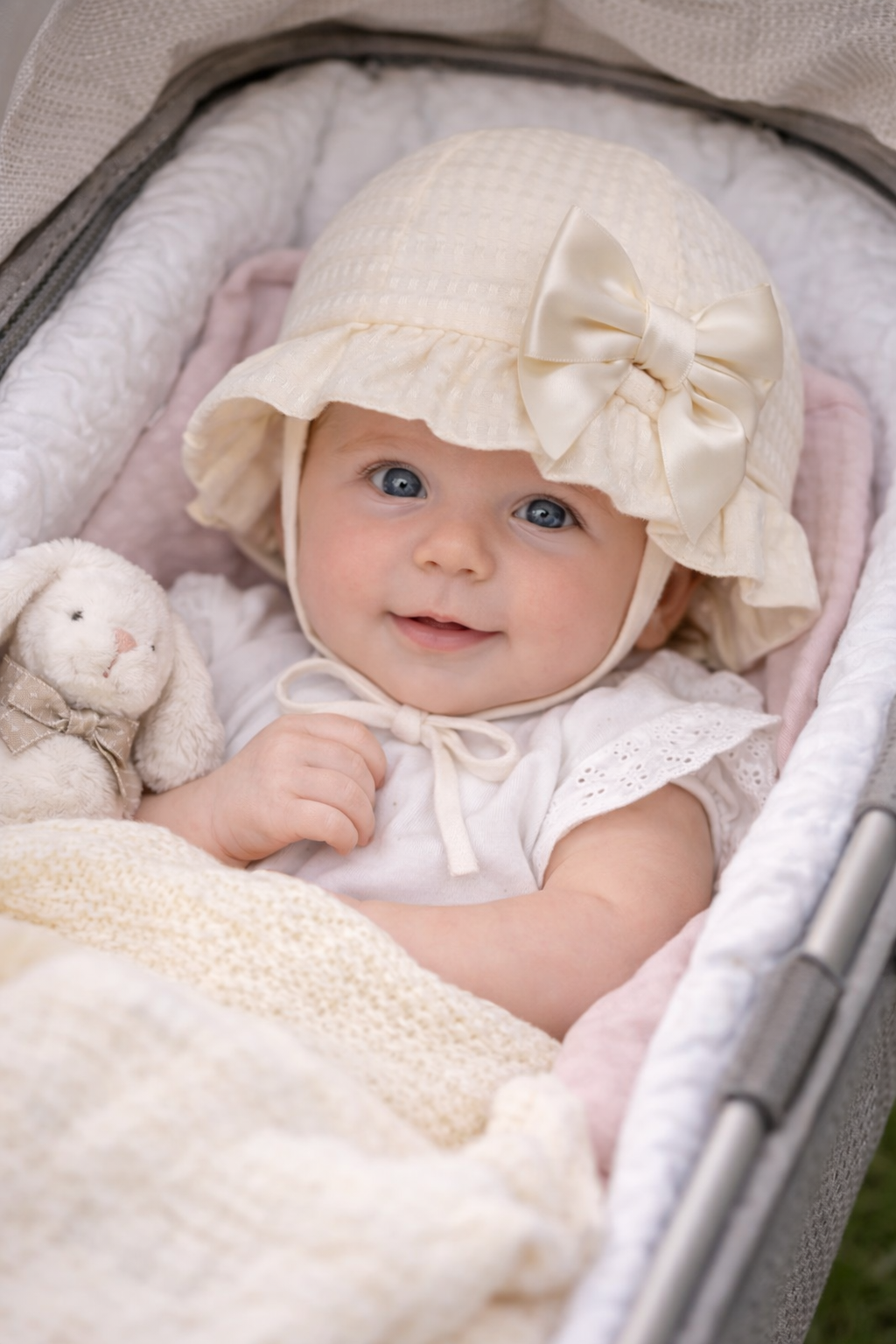 Baby in a pram wearing a white bonnet with a bow, surrounded by soft textures and toys.