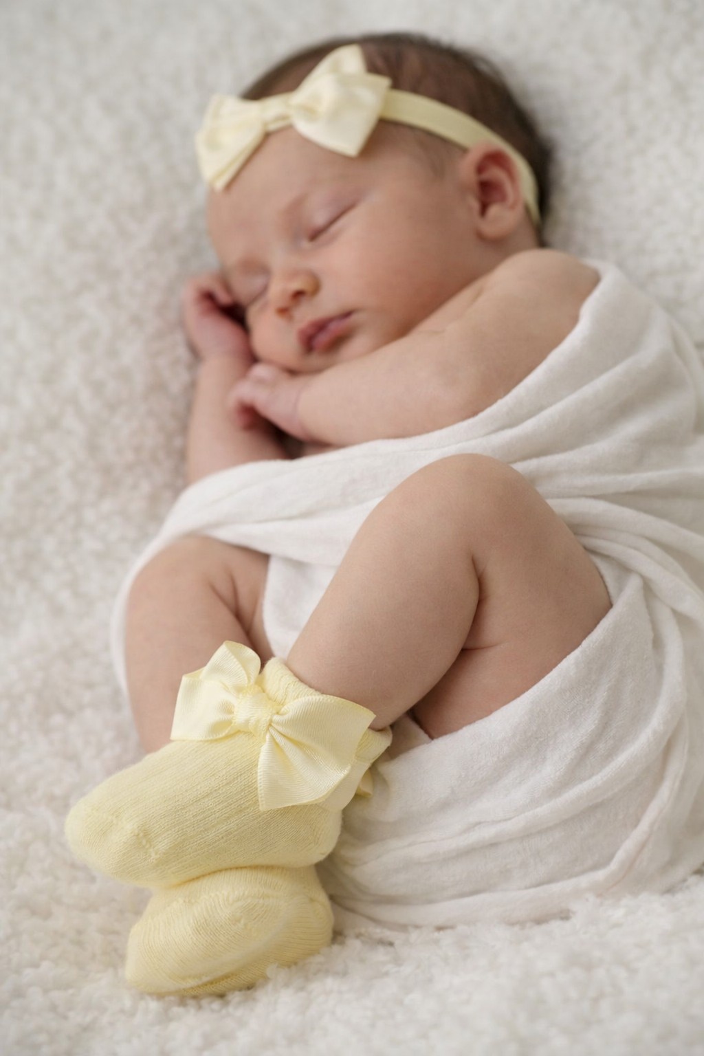Newborn baby wrapped in white with yellow booties and headband on a soft surface