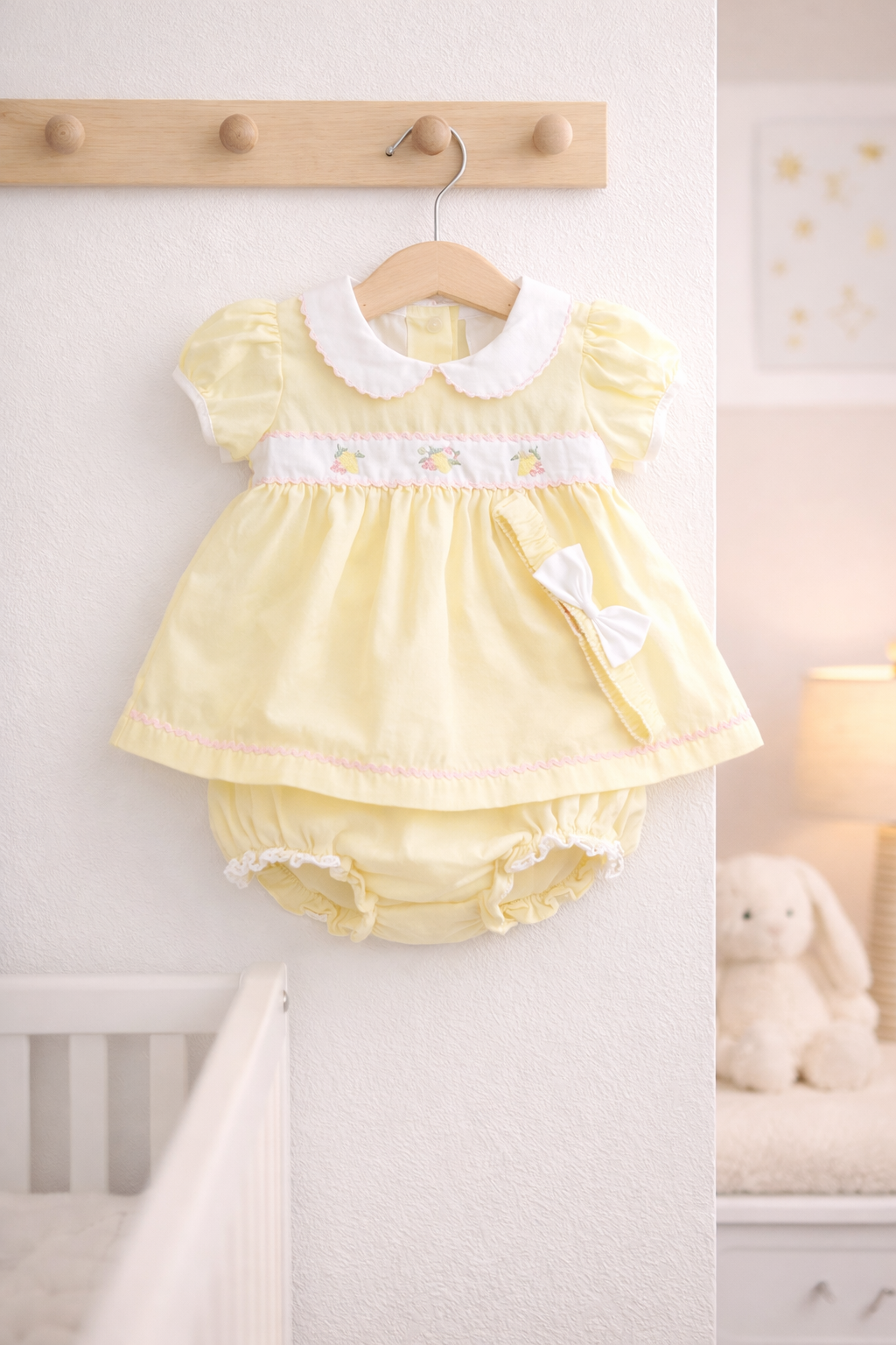 Yellow baby dress with white trim hanging on a wooden hook against a white wall.