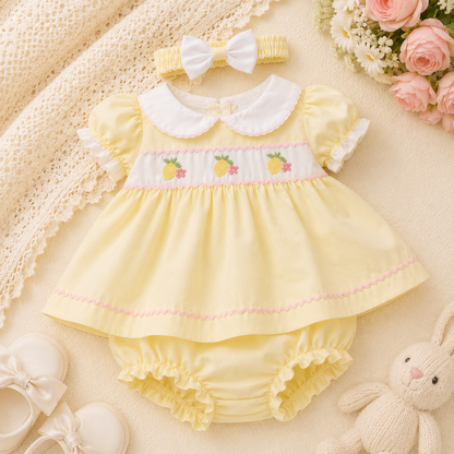 Yellow baby dress with white collar, matching headband, and bloomers on a soft surface with flowers and toys.