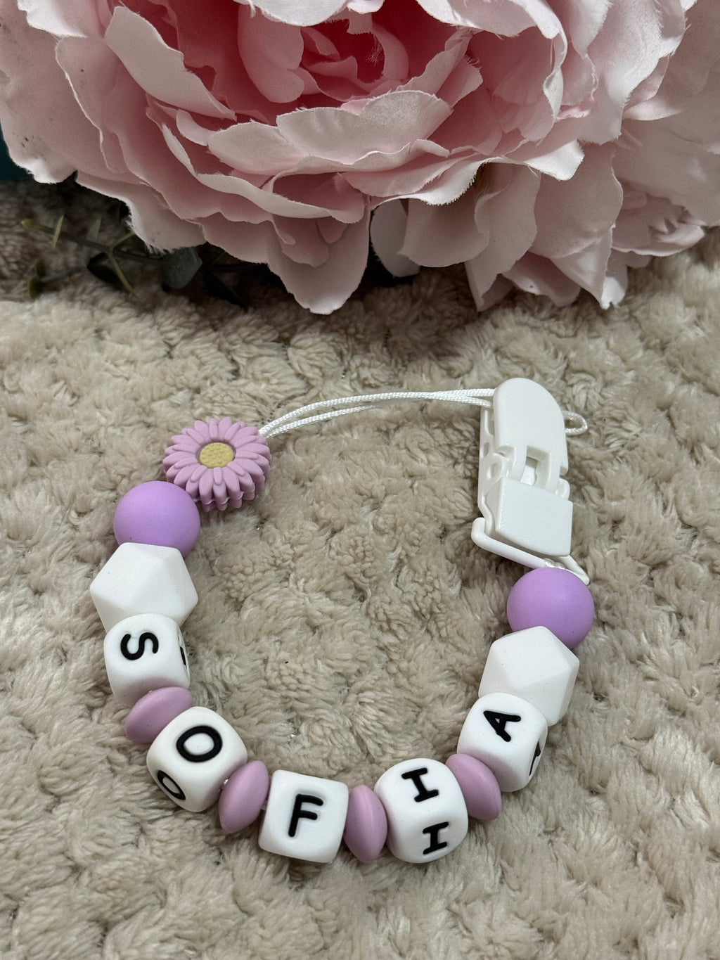 dummy clip with letter beads spelling 'Sofia' next to a pink flower on a textured surface