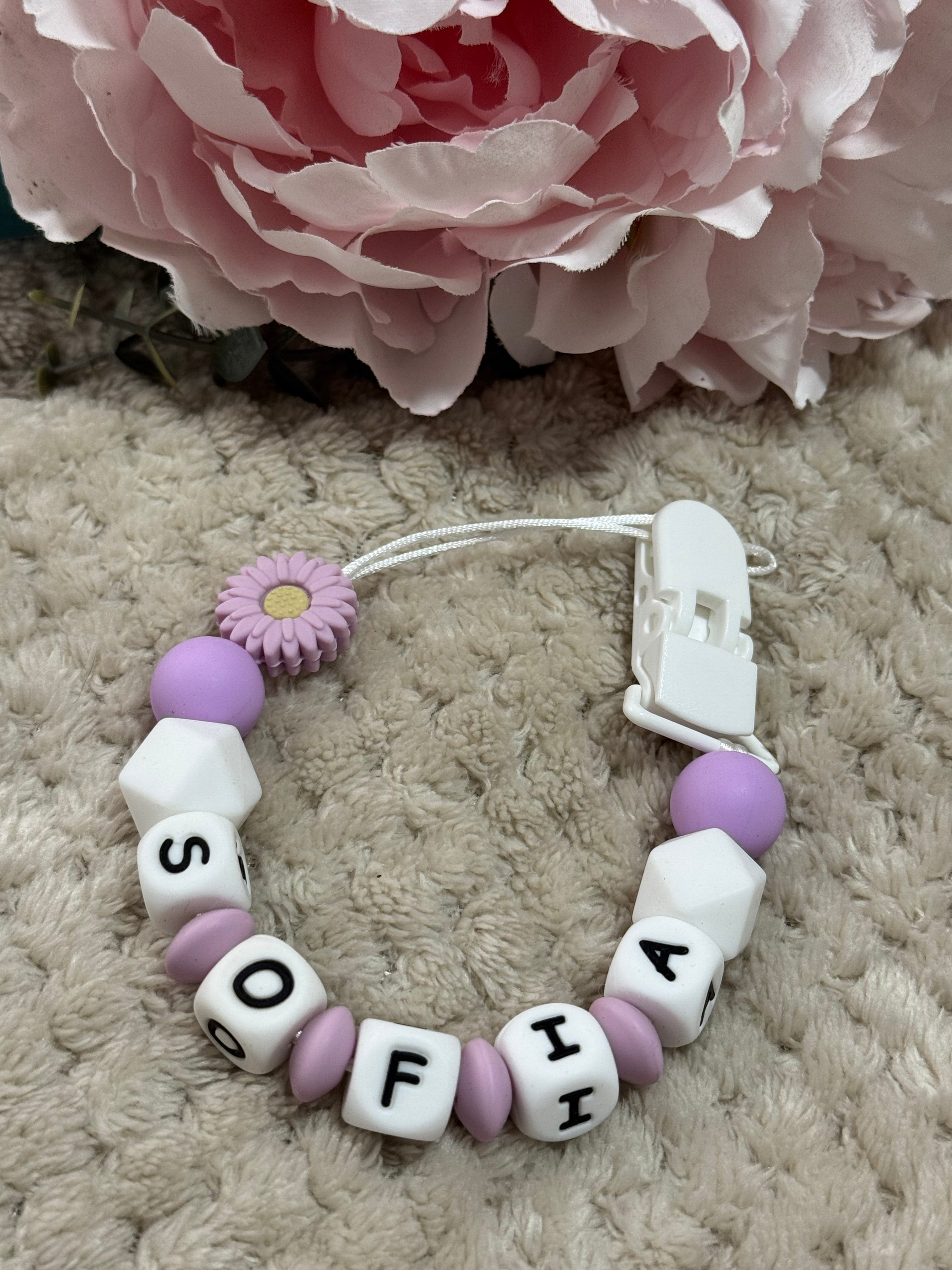 dummy clip with letter beads spelling 'Sofia' next to a pink flower on a textured surface