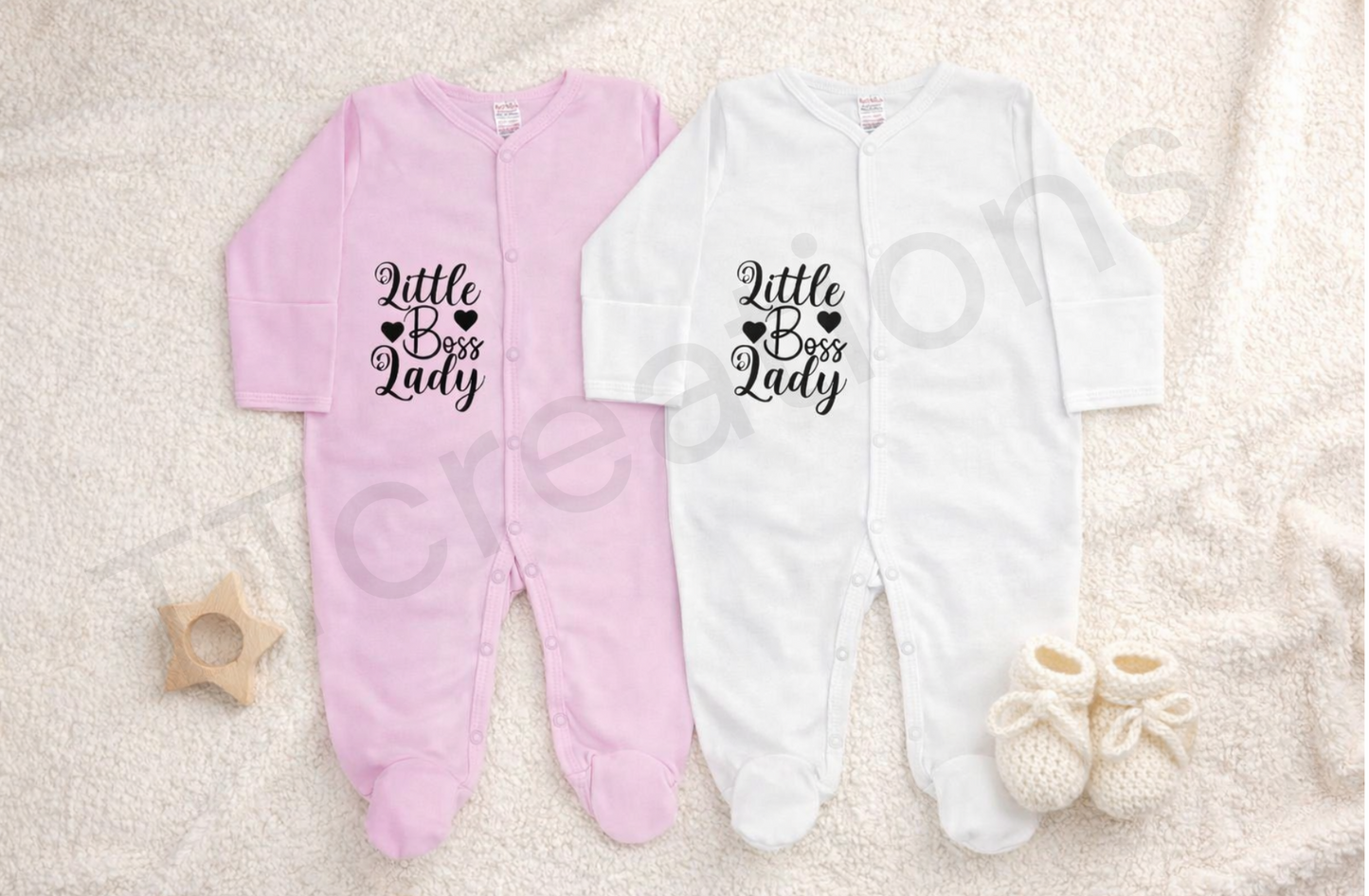 Pink and white baby footed sleepsuits displayed on a soft neutral blanket, featuring the phrase “Little Boss Lady” printed in black script with heart details. The newborn sleepsuits have long sleeves, snap button fastening, and soft cotton fabric, styled with knitted baby booties and a wooden star teether for a clean