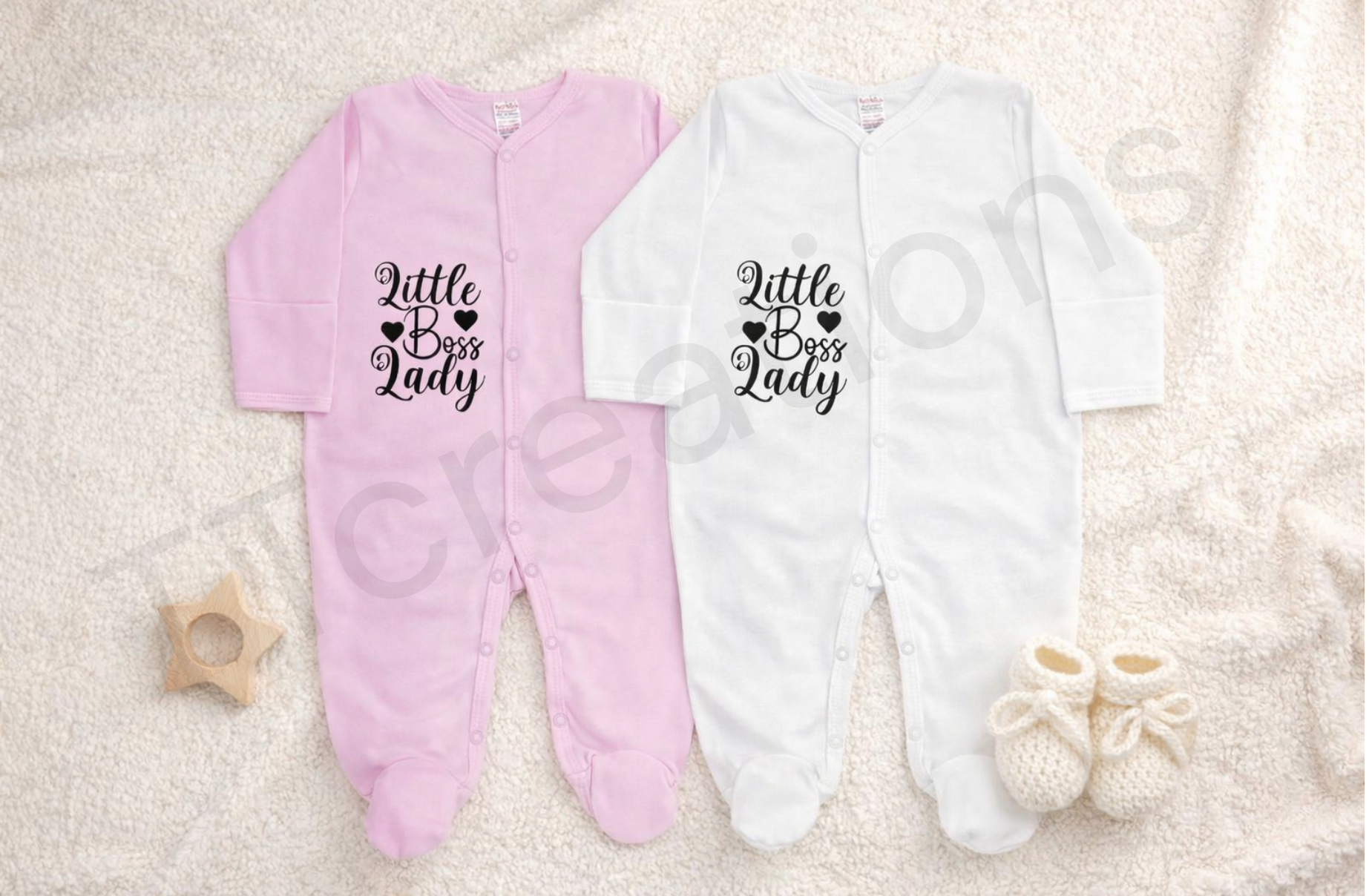 Pink and white baby footed sleepsuits displayed on a soft neutral blanket, featuring the phrase “Little Boss Lady” printed in black script with heart details. The newborn sleepsuits have long sleeves, snap button fastening, and soft cotton fabric, styled with knitted baby booties and a wooden star teether for a clean