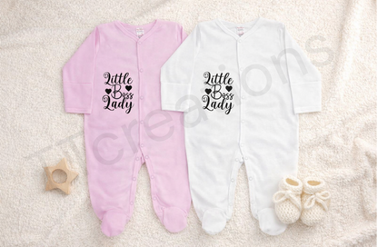 Pink and white baby footed sleepsuits displayed on a soft neutral blanket, featuring the phrase “Little Boss Lady” printed in black script with heart details. The newborn sleepsuits have long sleeves, snap button fastening, and soft cotton fabric, styled with knitted baby booties and a wooden star teether for a clean
