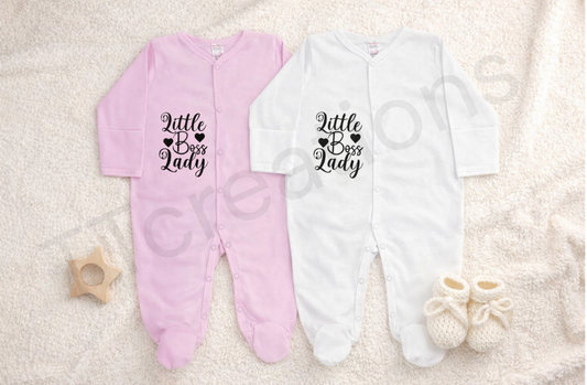 Pink and white baby footed sleepsuits displayed on a soft neutral blanket, featuring the phrase “Little Boss Lady” printed in black script with heart details. The newborn sleepsuits have long sleeves, snap button fastening, and soft cotton fabric, styled with knitted baby booties and a wooden star teether for a clean